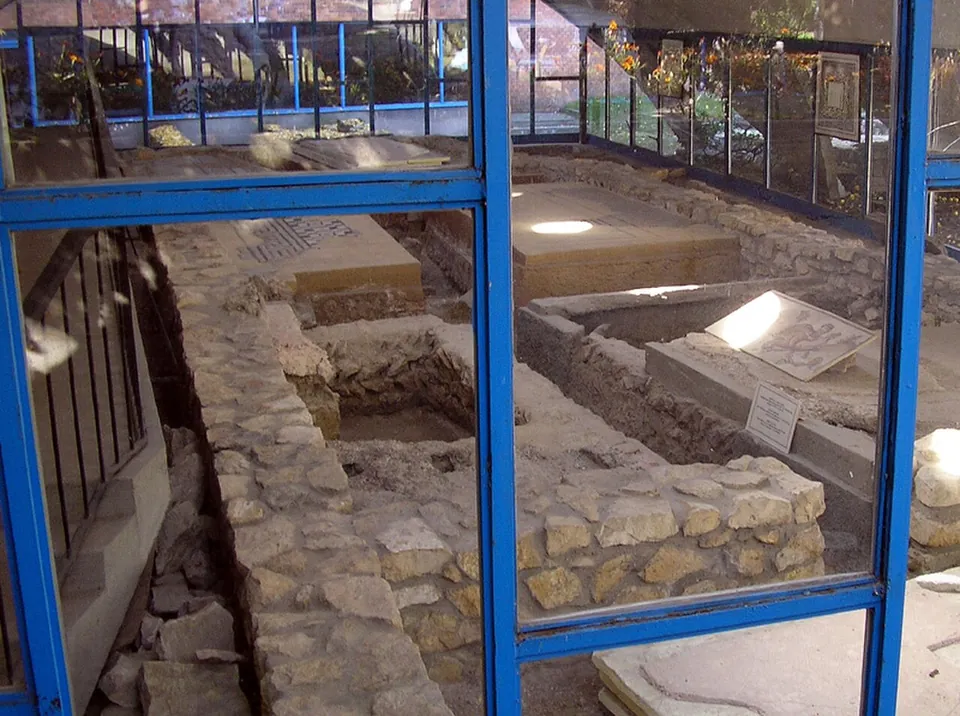 Seen through the blue-framed glass walls of a shelter, low stone walls of excavated rooms with patches of mosaic floor and small information labels set among them.