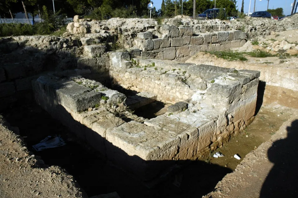 A sunken stretch of thick wall built of large squared limestone blocks, with rubble masonry above and parked cars and trees at the roadside behind.