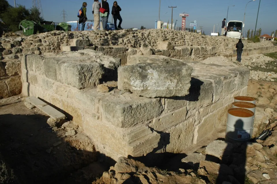 The squared stone blocks and rubble core of a ruined wall in an excavation trench, with a group of visitors standing on top and a coach and road signs behind.