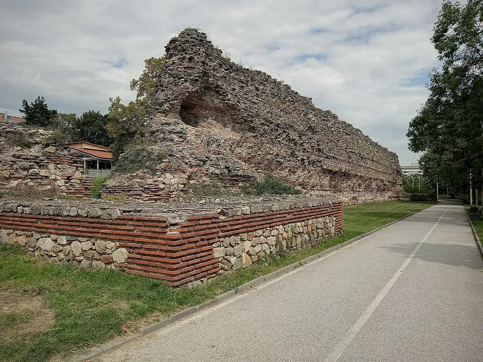 A tall, weathered stretch of city wall of rough stone with bands of brick, its top broken and ragged, beside a paved path and a low reconstructed wall with red brick courses in the foreground.