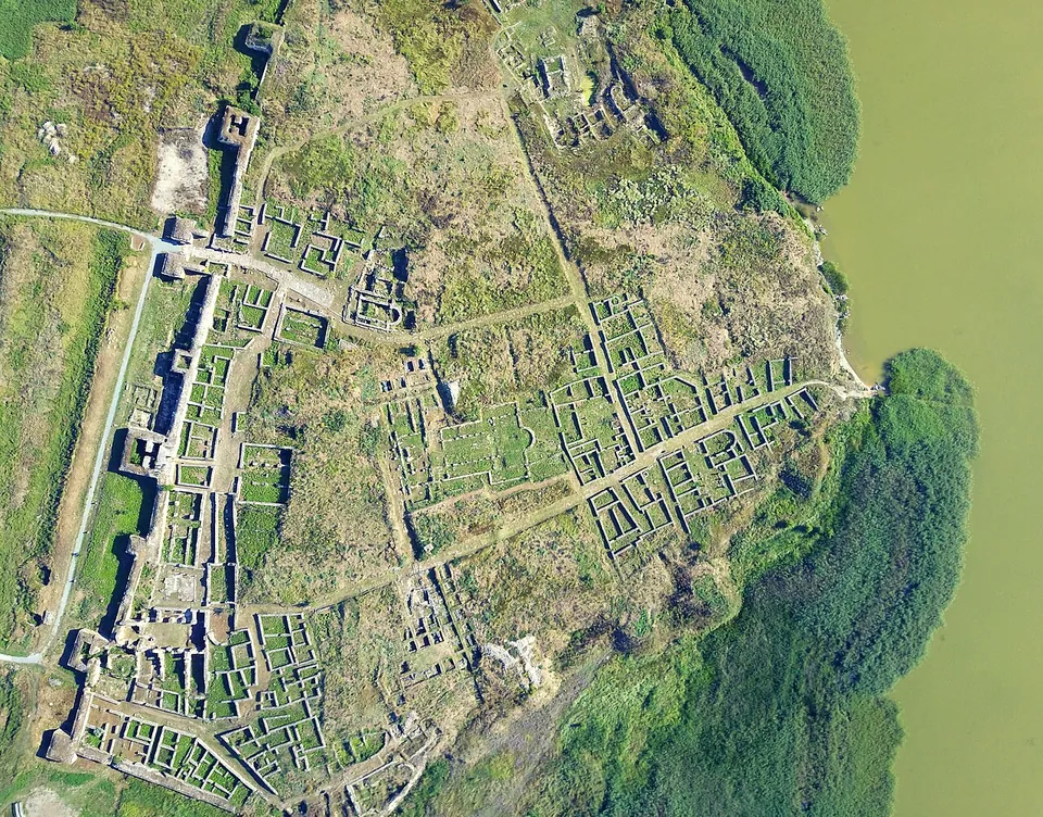 A vertical aerial view of a walled ancient city on a low point of land beside a green lake, the dense grid of excavated stone foundations and streets bounded on one side by a long wall with towers, and reed beds along the shore.