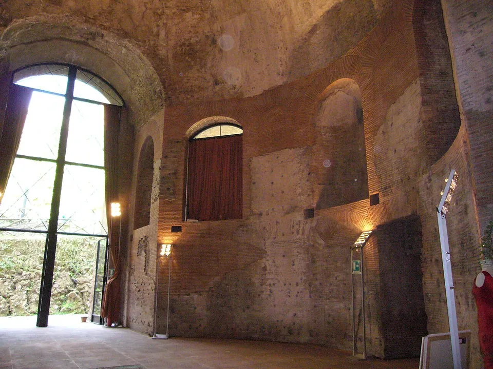 The inside of a tall brick hall lit by lamps, its walls set with round-headed niches and rising into a dome, with an open doorway through to a further room; dresses on stands and a small glass panel in the floor show it in use for an exhibition.