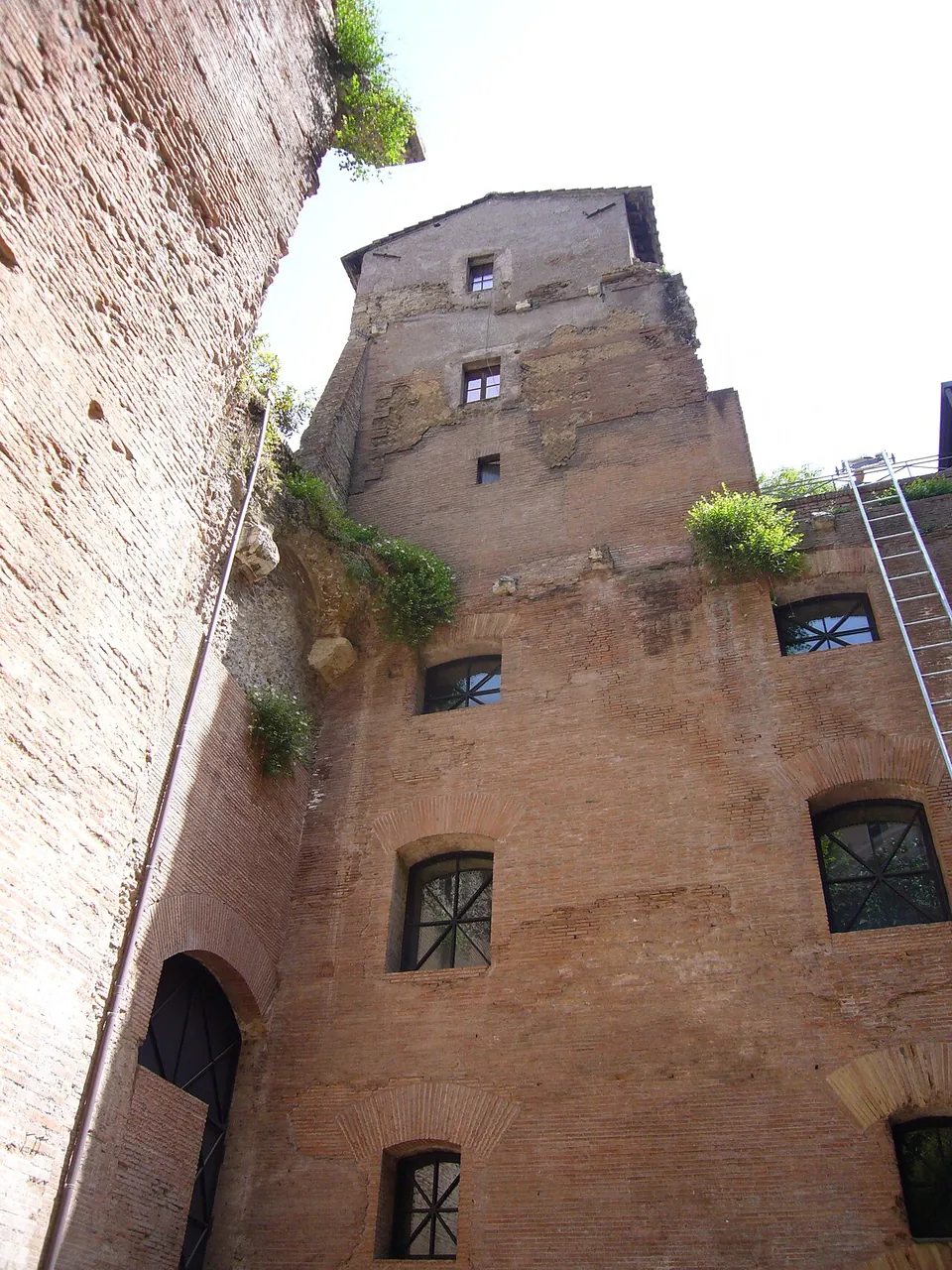 Looking up at a high Roman brick building with rows of windows on several floors, its top ragged, with a metal ladder against the wall and the brick side of the hall at the left.