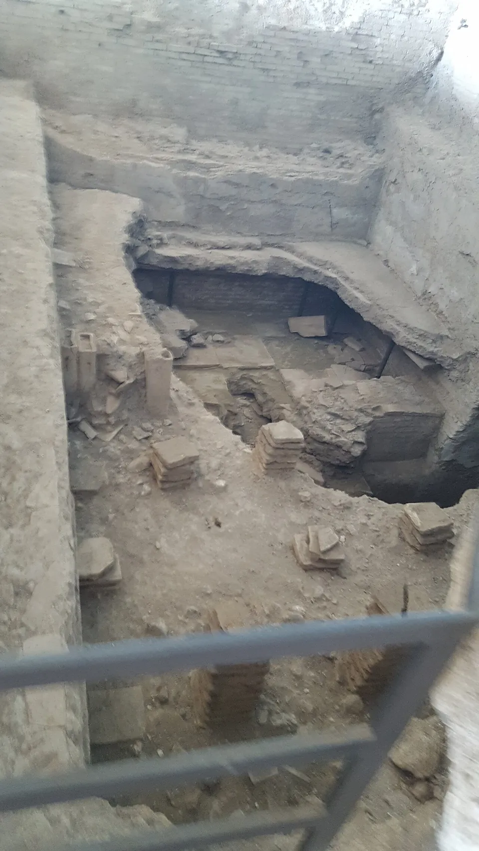 Looking down past a metal railing into an excavated pit with brick walls and the stubs of small brick stacks on its floor.