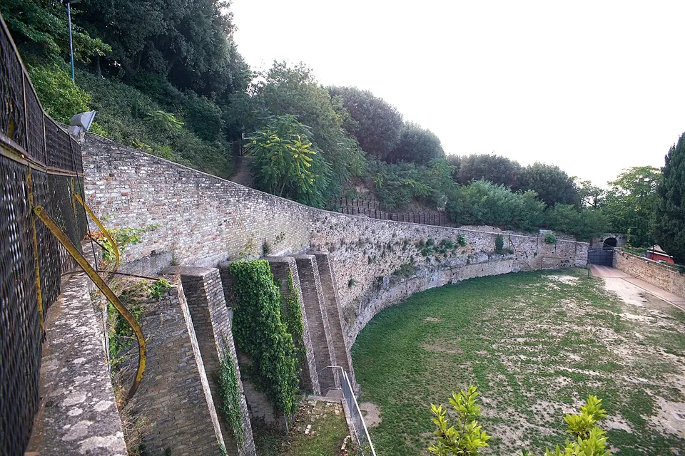 A long curving wall of brick and stone with buttress-like piers below a wooded slope, bordering an open grassy area, with a railing along the top.