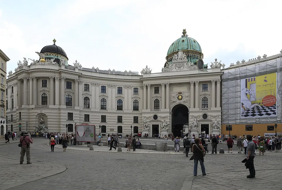 The domed Michaeler wing of the Hofburg palace behind a cobbled square full of people, with a low walled enclosure in the middle of the square in front of the central gateway.
