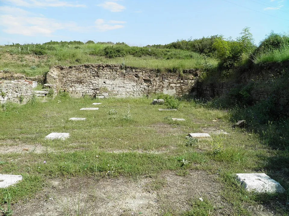 A wall of stone and brick at the back of a grassy space, with rows of square marble bases set in the ground in front of it.