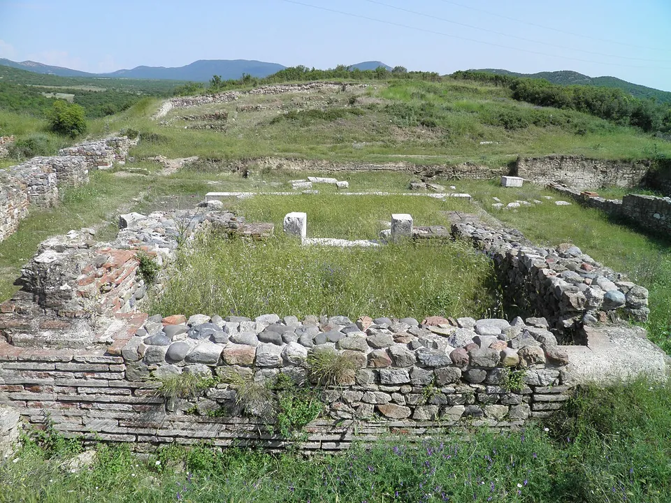 Rooms of an excavated building with walls of stone bonded with courses of red brick, two small marble pillars standing inside, and grassy hills and mountains behind.