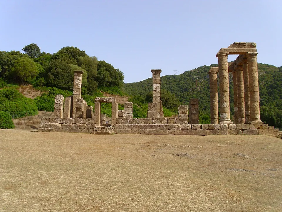 The temple seen across a wide, dry, sandy clearing, its long low platform of squared stone running from left to right, a few pillars and wall stubs standing on it and the tall columns of the porch at the right end, with wooded hills behind.