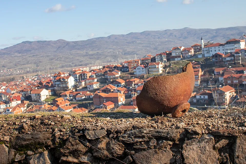 A large broken clay vessel lying on its side on top of a stone wall, with the houses of the town and a hill range behind.