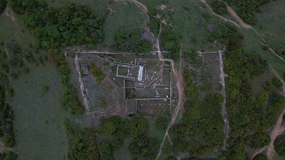 An aerial view of a rectangular walled enclosure on a hilltop, with the outlines of buildings inside it, surrounded by scrub, trees and dirt tracks.