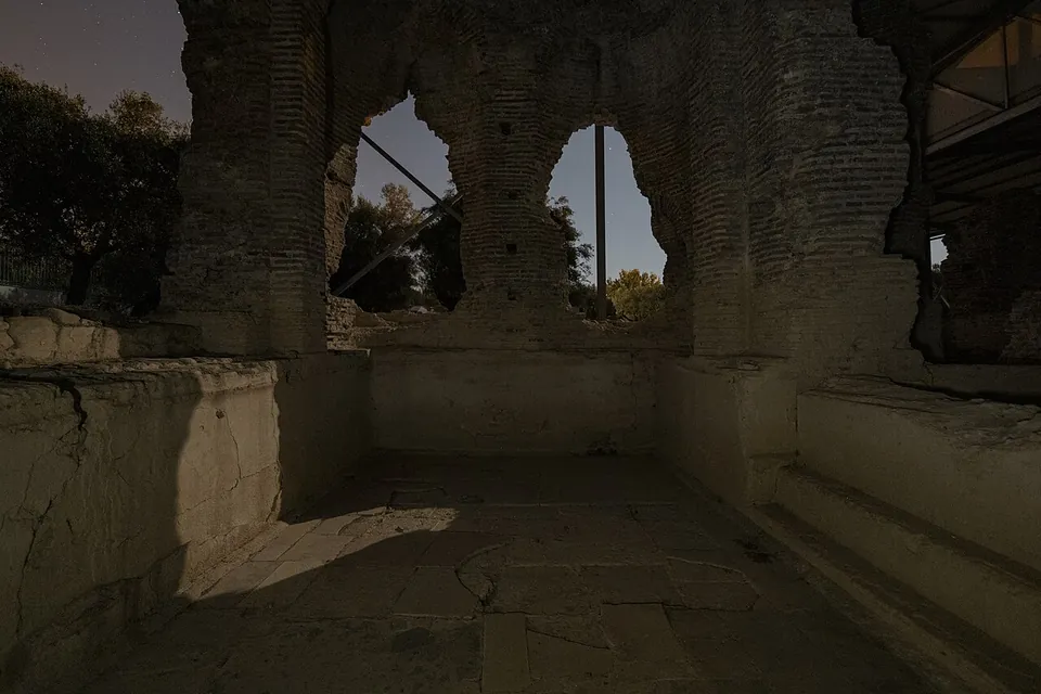 Tall ruined walls of brick with two arched window openings, above a sunken basin with a paved floor and a step along one side, lit at night under a protective roof.