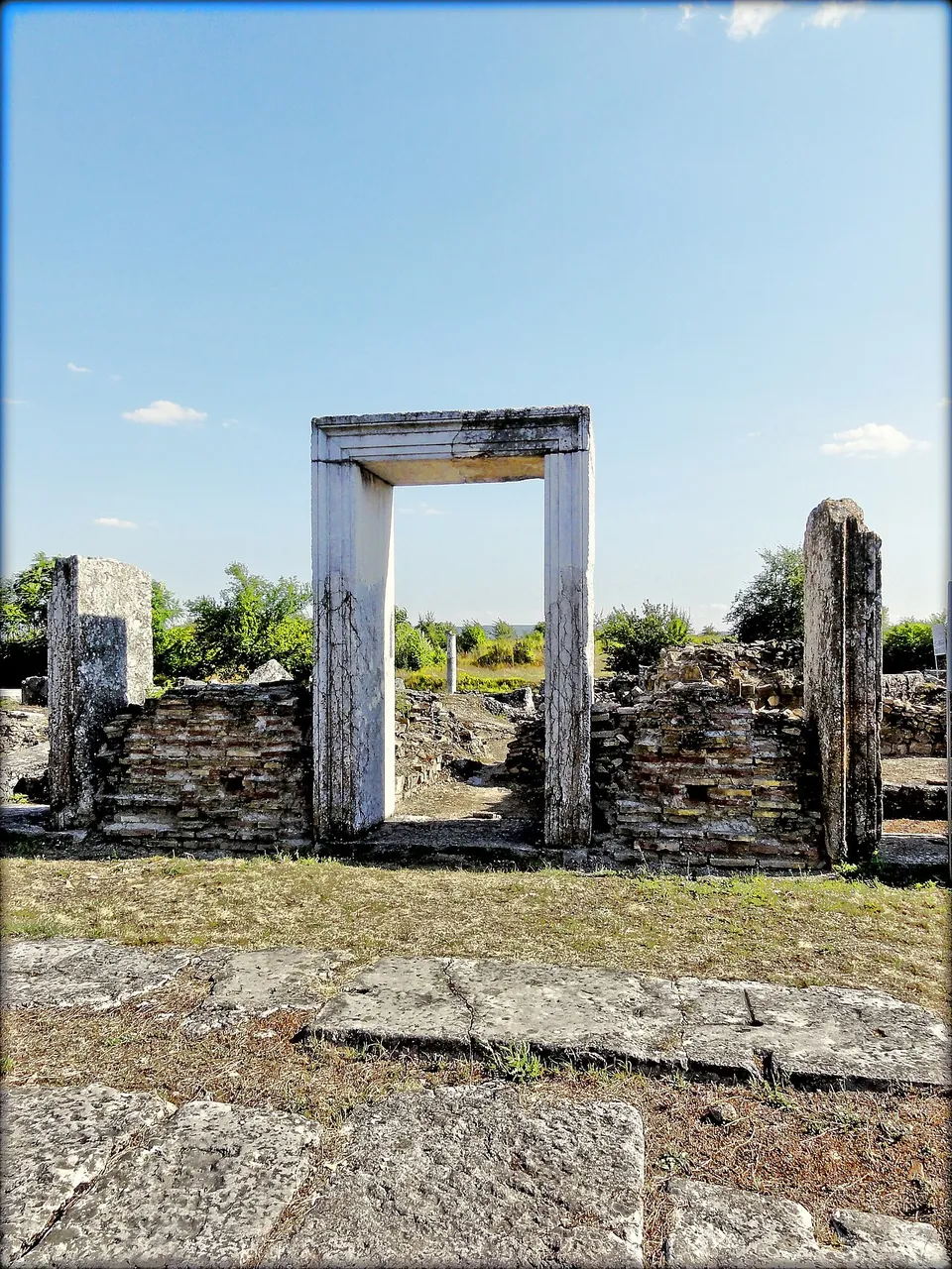 A re-erected marble doorframe standing alone in a low wall of brick and stone, with a stone-paved street in the foreground and greenery beyond.