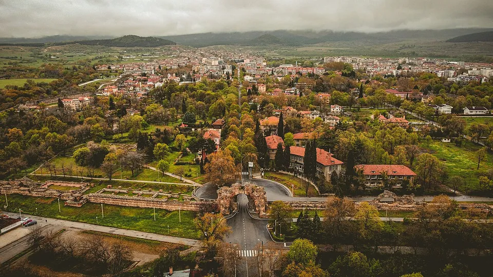 An aerial view over the spa town of Hisarya, its red roofs and trees spreading to the hills, with a road running through a ruined Roman gateway in the foreground and the low line of the city wall stretching away to the left across lawns.