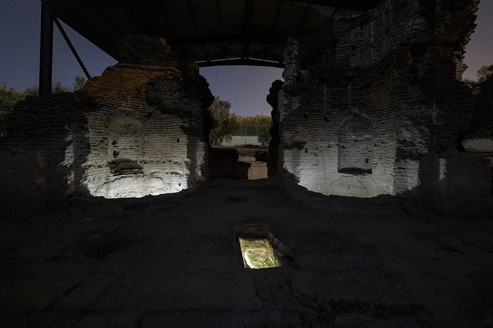 Two massive brick structures with niches in their walls, lit at night under a steel roof, with a small lit opening in the floor between them.