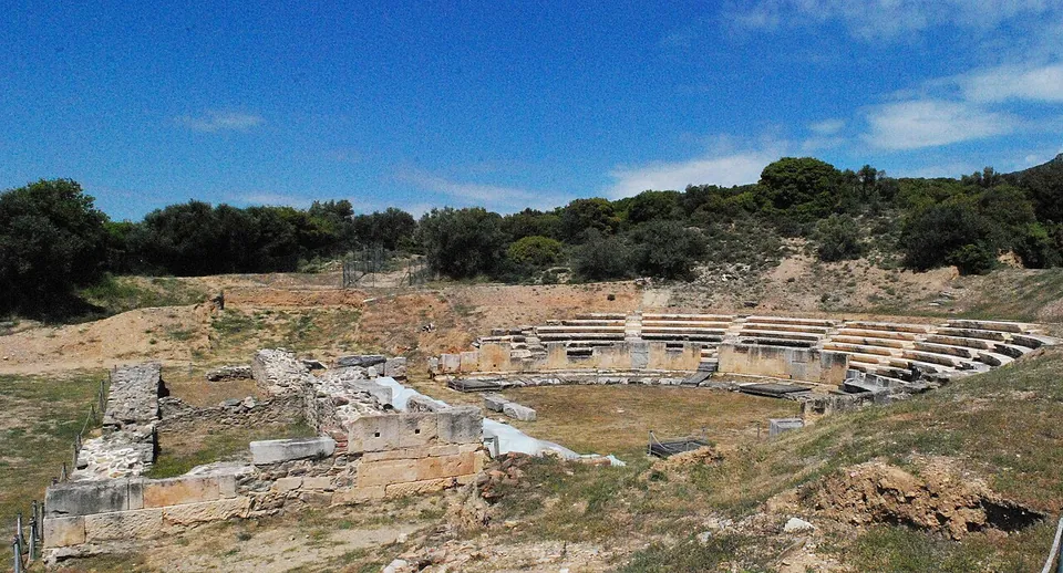 The restored lower rows of a stone theatre in a hollow below wooded hills, with ruined walls of the stage building in the foreground and a grassy orchestra between.