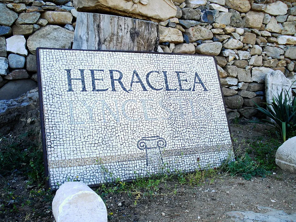 A white mosaic panel reading "Heraclea Lyncestis" in dark letters, with an Ionic capital beneath, propped against a dry-stone wall.