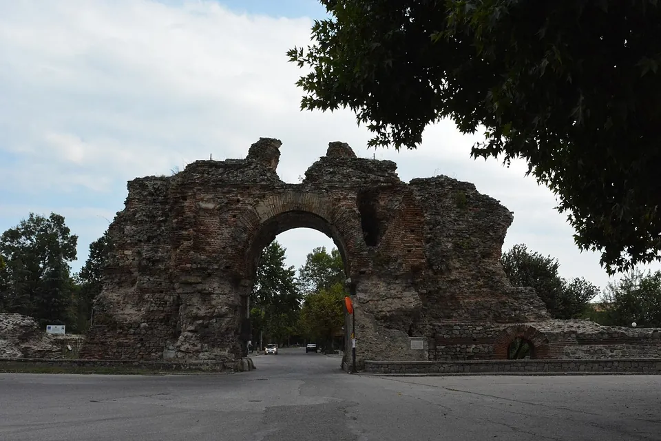 A massive ruined gateway of stone and brick, its central arch still standing between two broken masses of masonry that rise above it, with a road running through the arch and trees on either side.