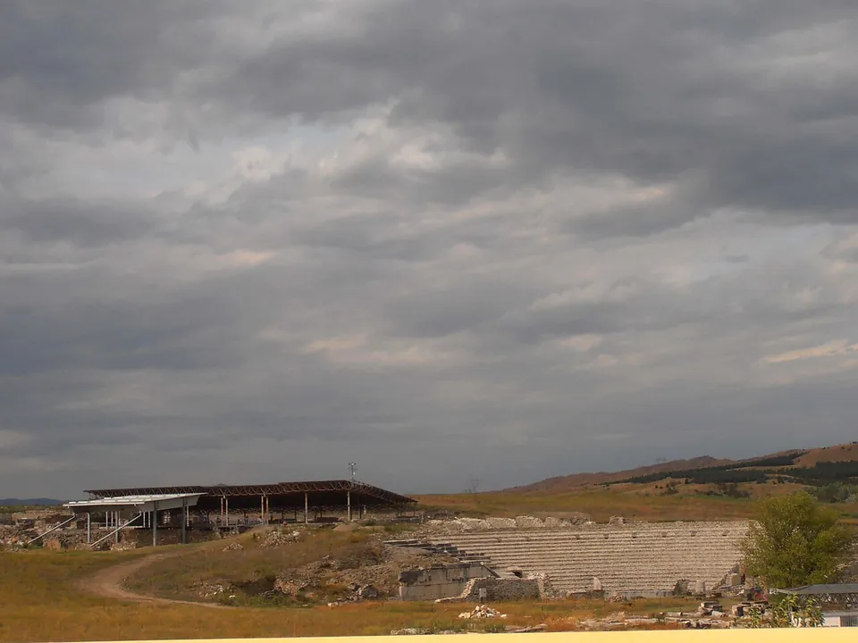 The curved stone seating of the theatre on a dry, open plain under a heavy grey sky, with a large modern shelter on stilts to the left and low hills behind.