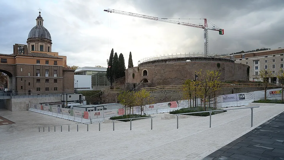 A wide, newly paved pale stone square with steel bollards and young trees in planters; beyond it a long run of white site hoarding encloses the round brick mausoleum, with a few cypresses on top and a tall construction crane above, a domed church and modern buildings at either side, under grey cloud.