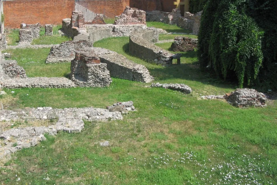 Low walls of rubble stone and brick in mown grass, among them a curving corridor around a round central space, with the brick wall of a modern building behind.