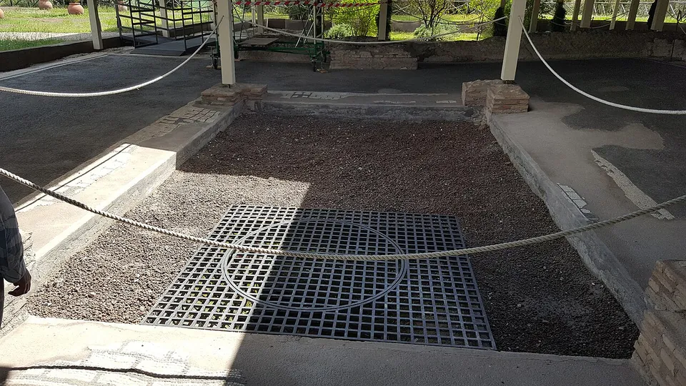 A sunken rectangular bed of gravel with a metal grating at its centre, edged by low stone kerbs and a grey paved floor, under a modern shelter roof on slender posts, with rope barriers keeping visitors back.