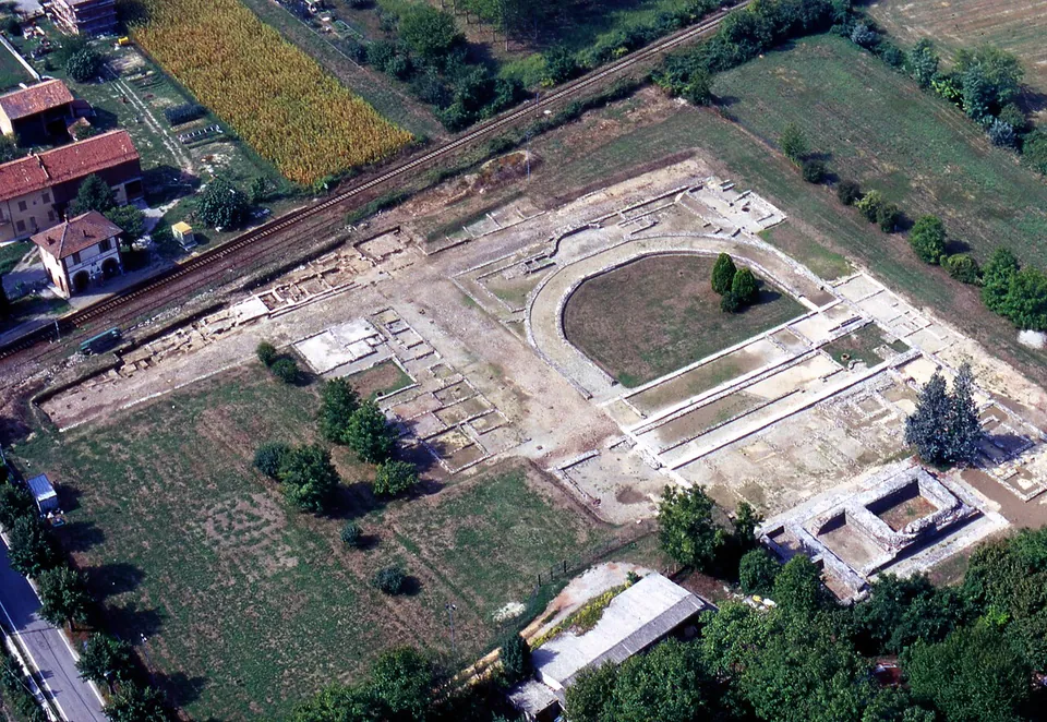 An aerial view of a large excavated rectangle of pale ground among fields and trees, laid out with low wall lines; in the middle a horseshoe-shaped enclosure with a lawn inside and, beside it, a rectangular walled structure, with a railway line and houses at the upper left.