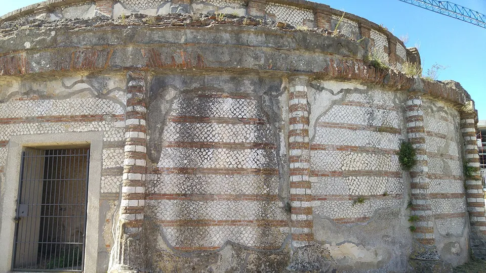 The curved outer wall of the tomb seen close up, faced with bands of small diamond-set stones between courses of red brick and divided by brick-banded pilasters, with a doorway closed by an iron grille at the left and plants growing from the top.