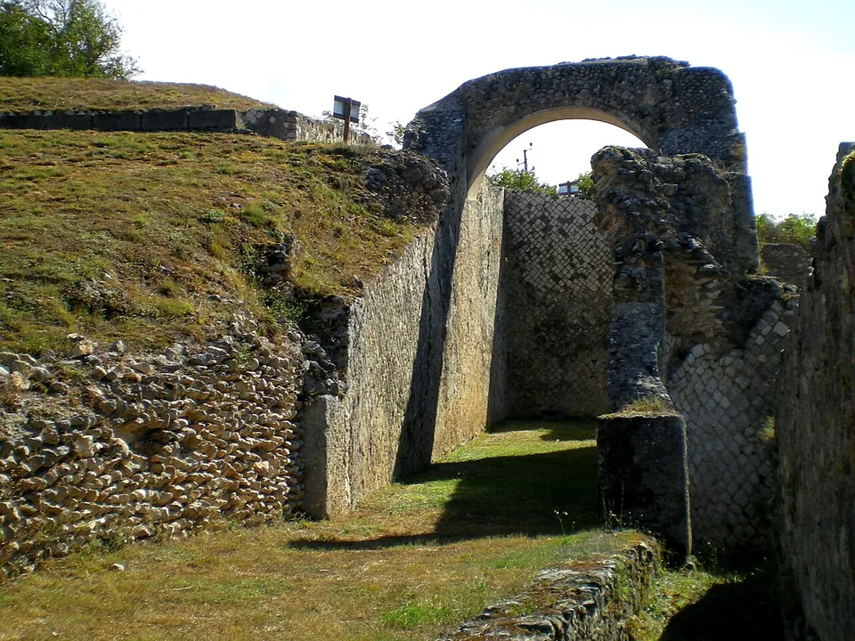 A stone arch spanning a grassy passage between high walls, one of them faced with small square stones laid in a diagonal net pattern.