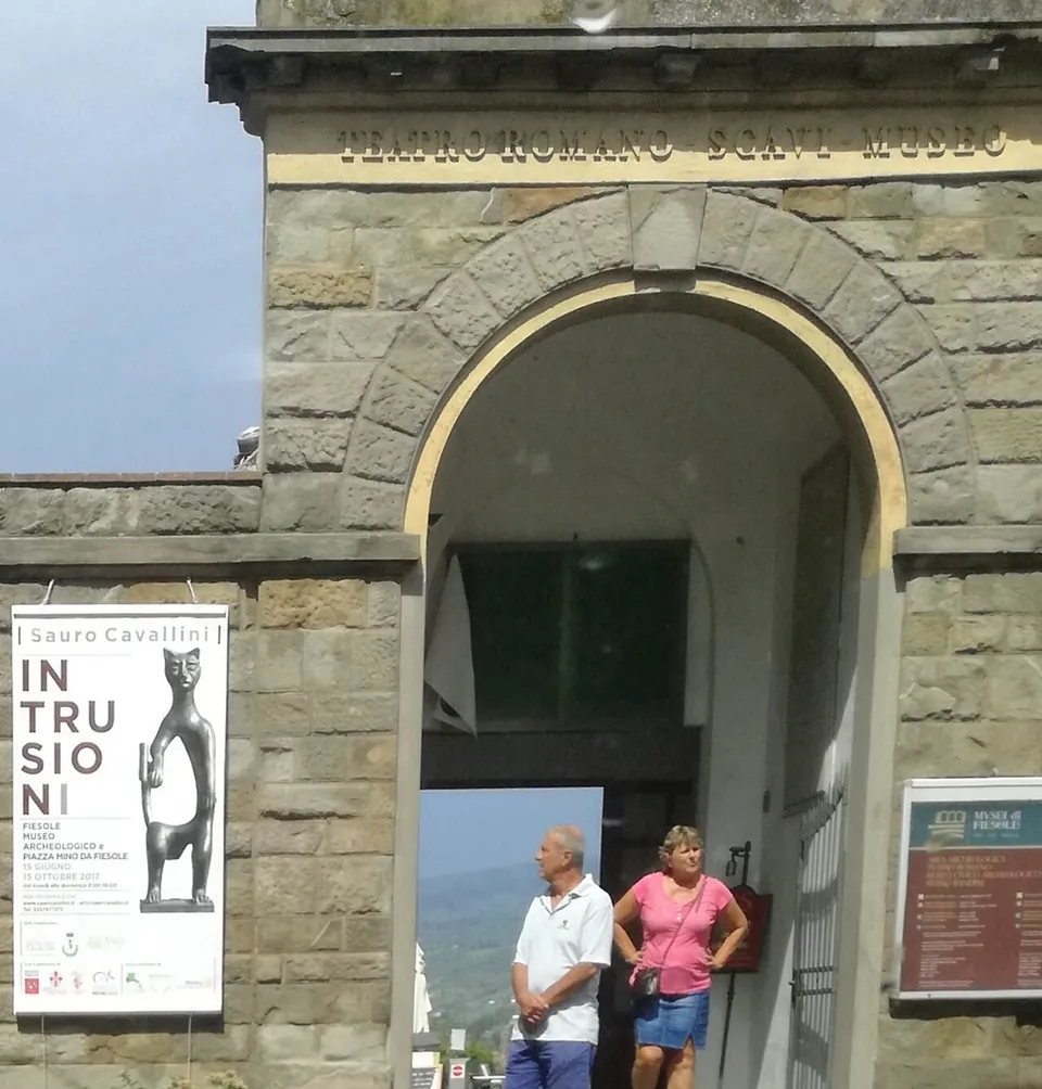 A stone gateway with a round arch, lettered "Teatro romano scavi museo" above it, a Musei di Fiesole information board on the right and an exhibition poster on the left, with two visitors standing in the opening.
