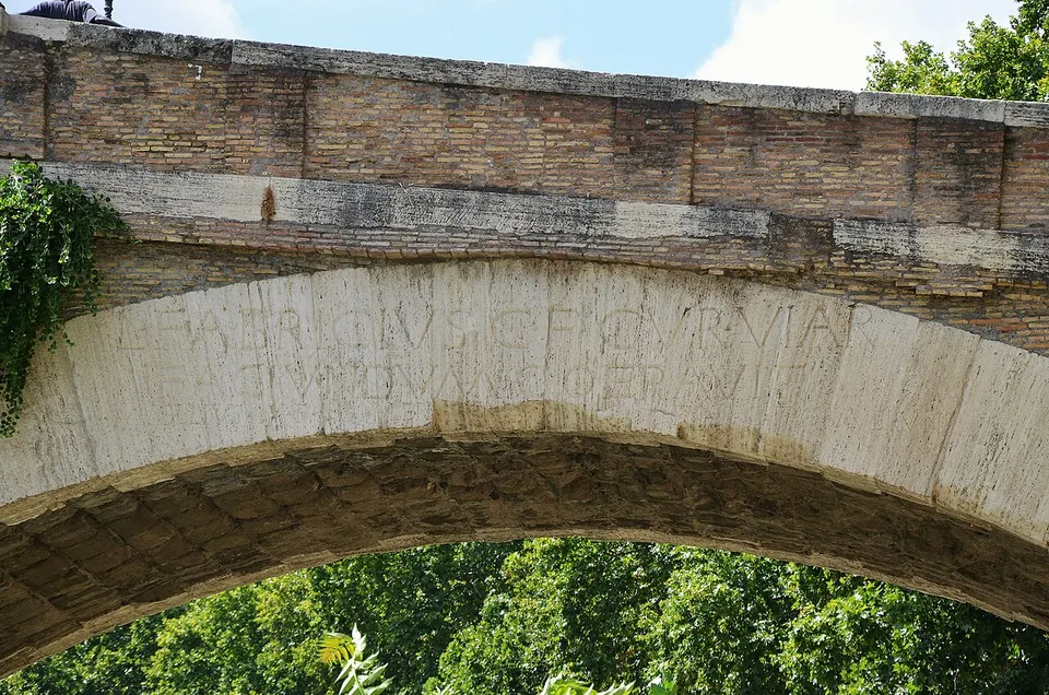 Large Roman capital letters cut into the pale stone blocks of a bridge arch, under courses of brick, with green trees seen through the arch below.