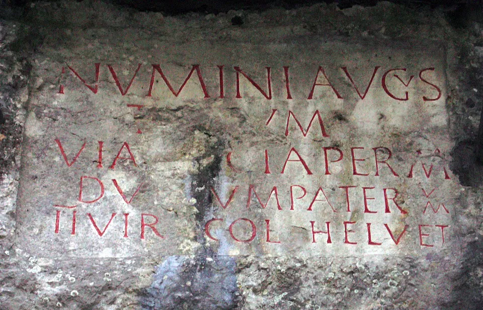 Roman capital letters cut into a smoothed panel of rough grey rock, picked out in red paint, some lines broken where the stone has flaked.