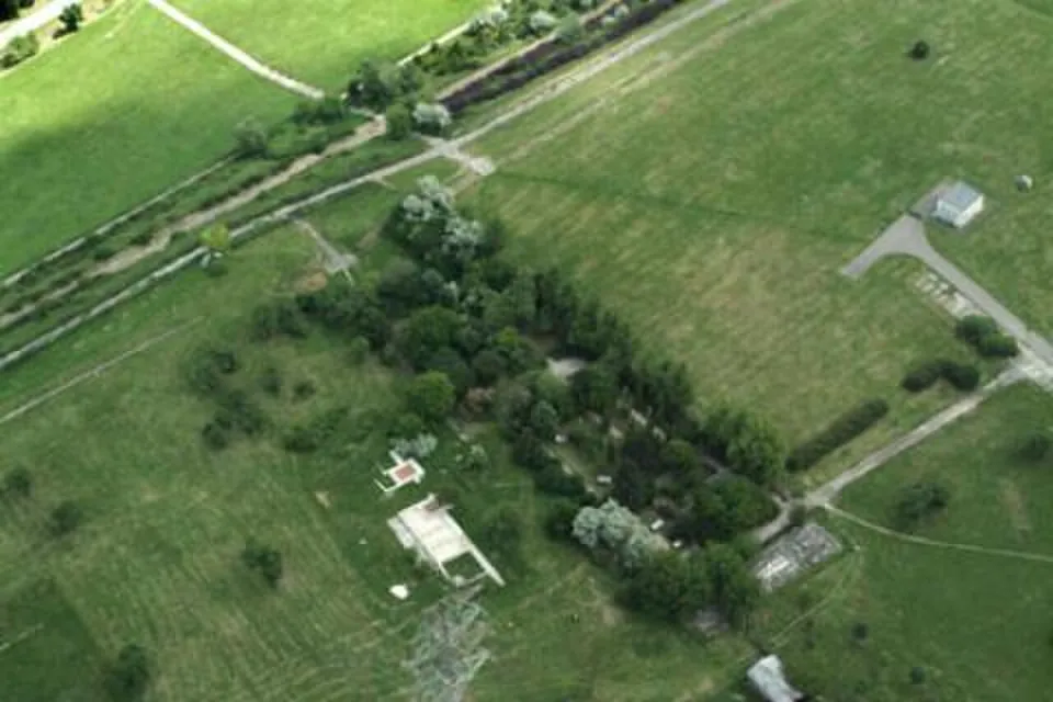 An aerial view of a grassy plateau crossed by paths and lines of trees, with a small white roofed structure and a pale rectangle of foundations among the grass and a power pylon in the foreground.