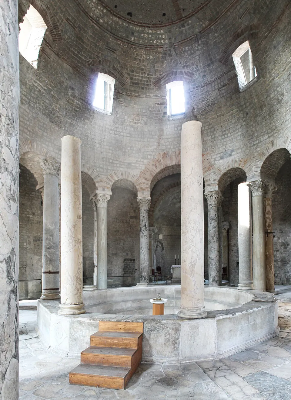 Inside a domed round hall, a wide circular font with a low marble rim and wooden steps up to it, ringed by tall plain columns, with paired columns and arches opening onto an ambulatory behind and round-headed windows high in the dome.