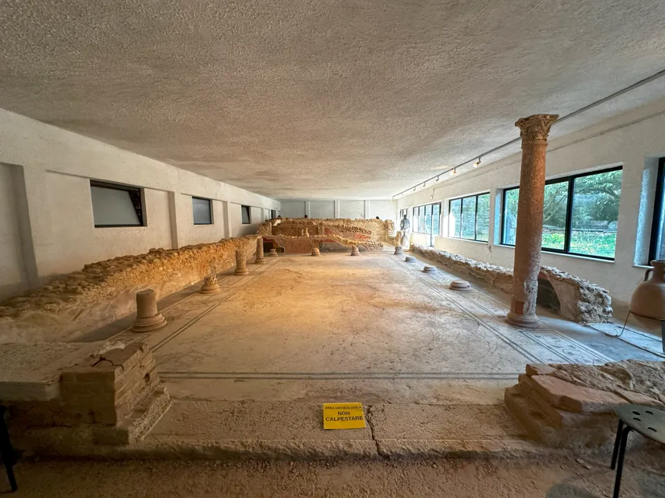 A long, low room under a flat modern ceiling, with large windows down the right-hand side. Low rubble walls and a row of short round column bases run along the edges of a pale mosaic floor edged with dark lines, and one column of pinkish stone with a carved capital stands to full height near the windows.