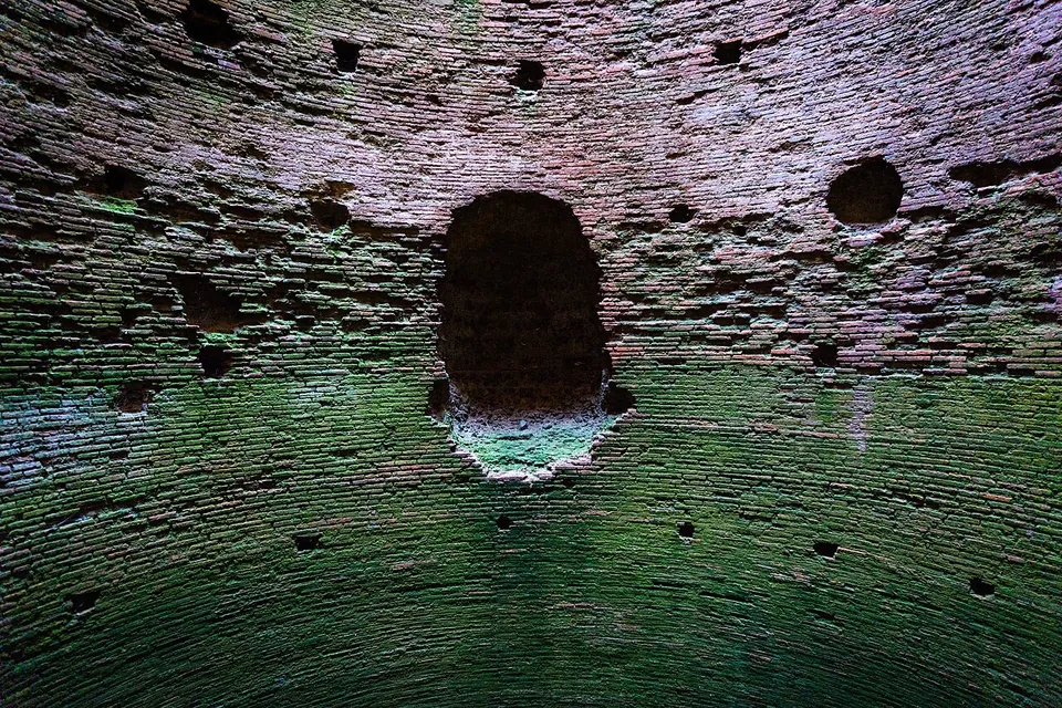 The inside of a round chamber of small brick-like stones, its curving wall pitted with rows of square holes and an arched opening, the lower courses stained green.