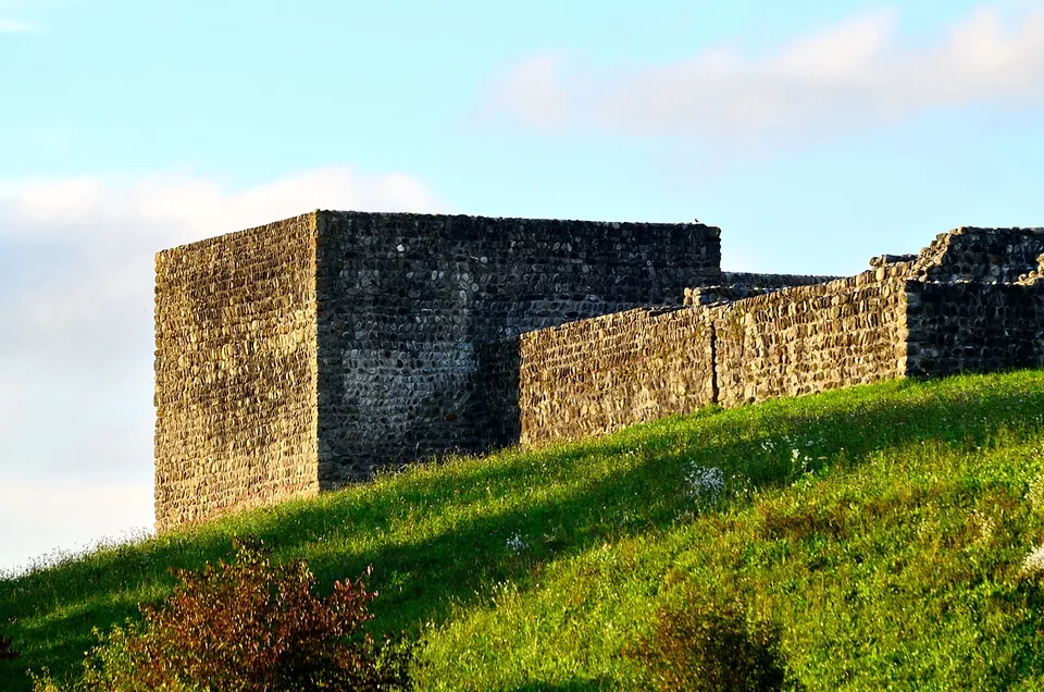 A tall square corner tower of rough stone and a long curtain wall running along the top of a grassy slope, lit by low evening sun.