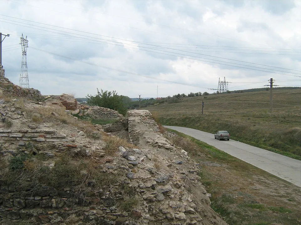 High ruined walls of rough stone with bands of brick on a bank to the left, beside a narrow paved road with a car on it, and pylons and power lines over green fields.
