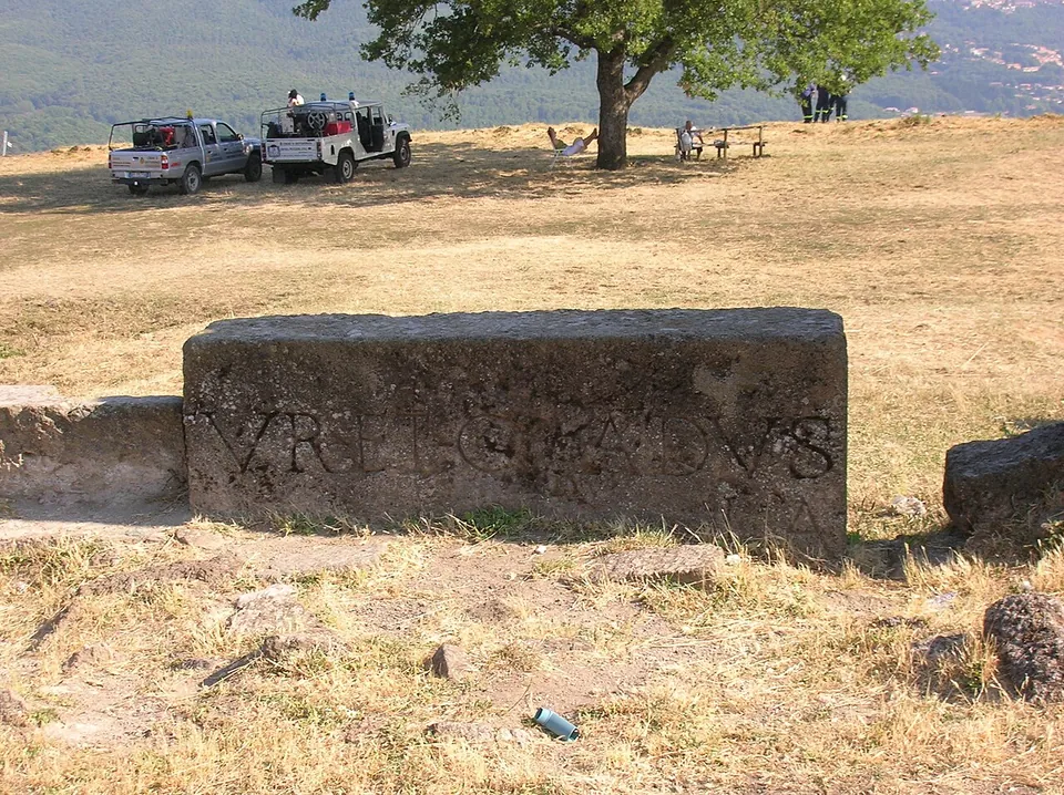 A long squared stone block lying in dry grass, its face carved with a line of large worn capital letters, with parked cars and people under a tree in the background.