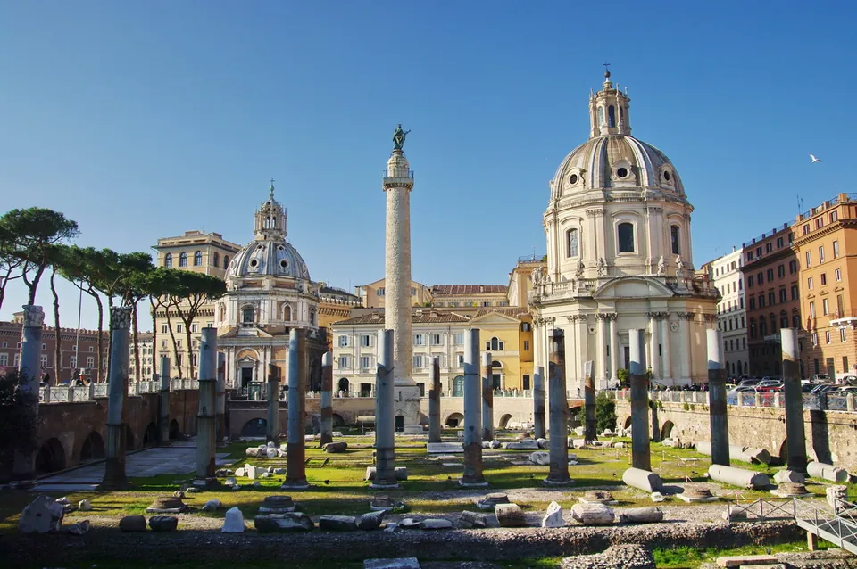 A lawn scattered with stone fragments and grey column shafts of differing heights, with a tall carved column topped by a statue standing between two domed churches beyond.