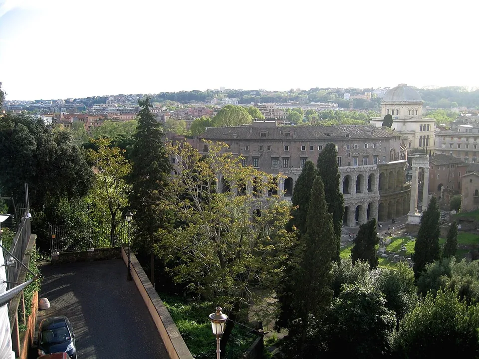 A view from a height over trees and a narrow lane to the curved arcades of the theatre, a tall building rising on top of them, with two free-standing columns to its right and the roofs of the city beyond.