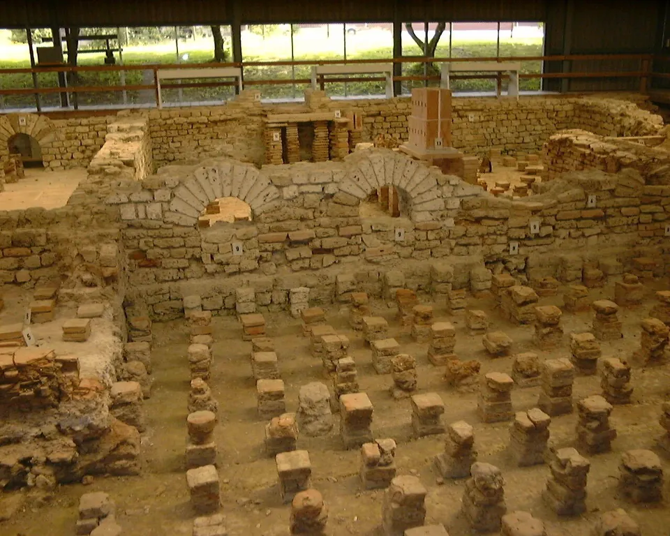 Rows of short square brick pillars standing on a floor between low stone walls, with two brick arches set into the wall behind, under a roofed shelter with windows.