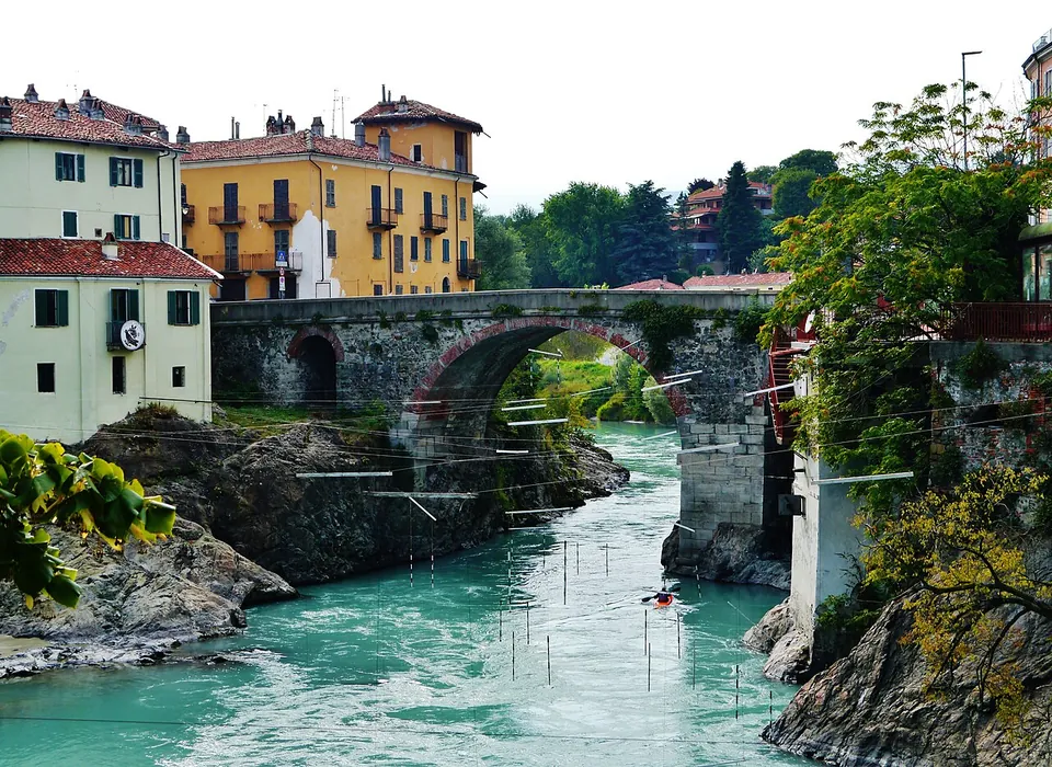 A stone bridge with one wide round arch spanning a fast turquoise river between rocky banks, poles hanging on wires above the water and tall yellow and white houses on the far bank.