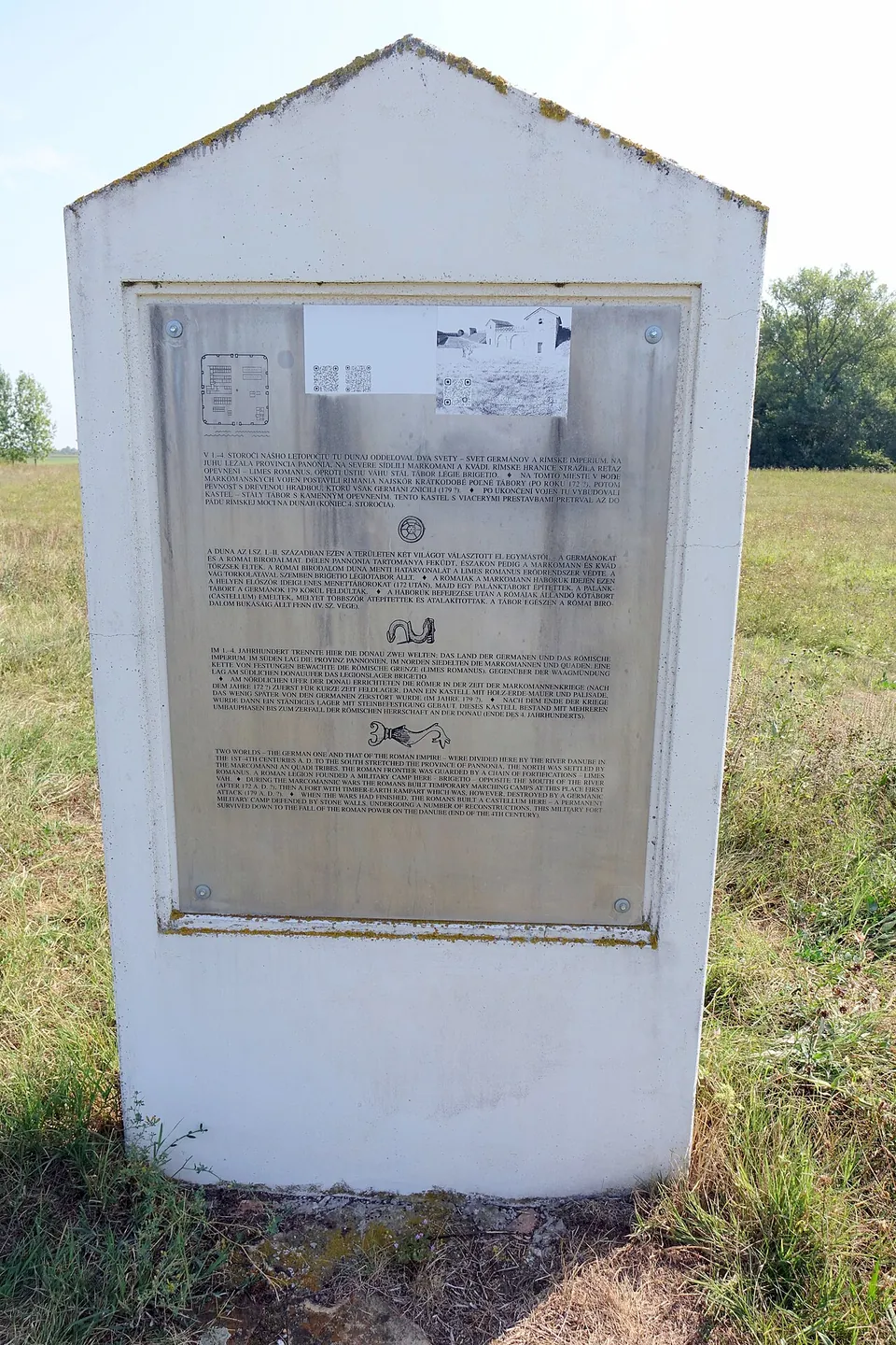 A white concrete stele in a meadow carrying a metal plate with a small plan of the fort and four blocks of text, in Slovak, Hungarian, German and English.