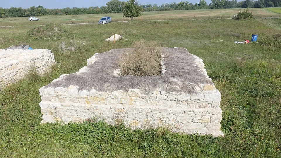 A rectangular block of neatly coursed pale stone, its top finished with mortar around a grassy hollow; cars stand on a track across the field behind.