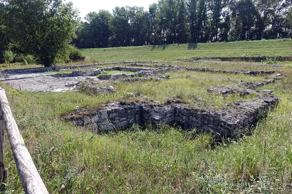 Low walls of rough grey stone laid out in rectangles in long grass behind a wooden rail, with a grassy bank and a line of trees along the back.