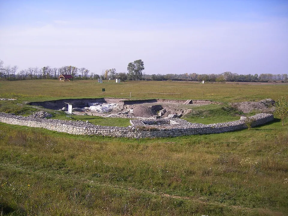 A flat meadow under a pale sky, with a long, curving low wall of rough stone in the foreground around an open excavation of earth heaps and pale stone; a small red-roofed hut and a line of trees stand on the horizon.