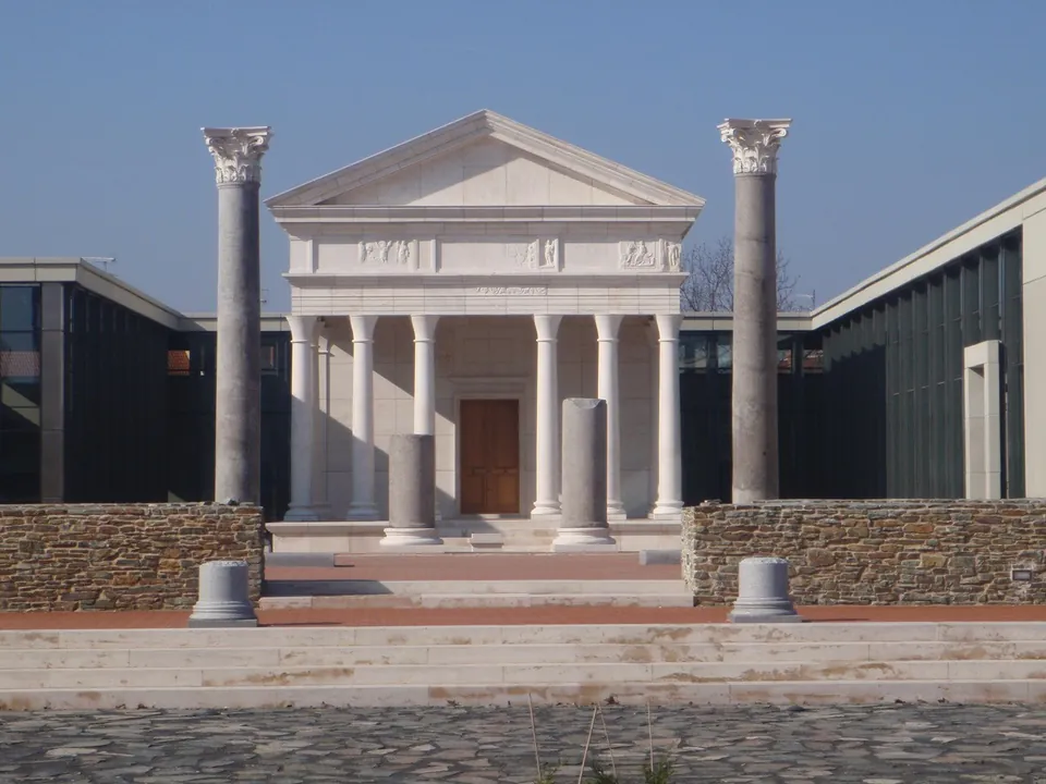 A white temple front with a triangular pediment on slender columns, set back behind two tall free-standing Corinthian columns and low walls of rough stone, with modern glass-and-concrete museum wings on either side.