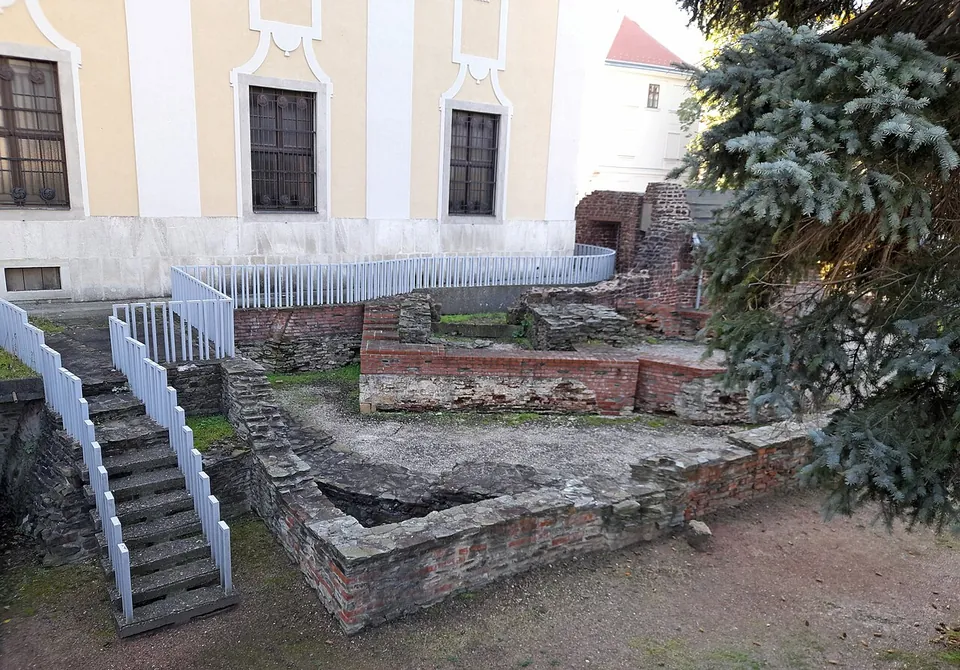 Excavated walls of brick and stone a little below street level, reached by a white-railed stair, beside a yellow building with ornate window frames and a fir tree.