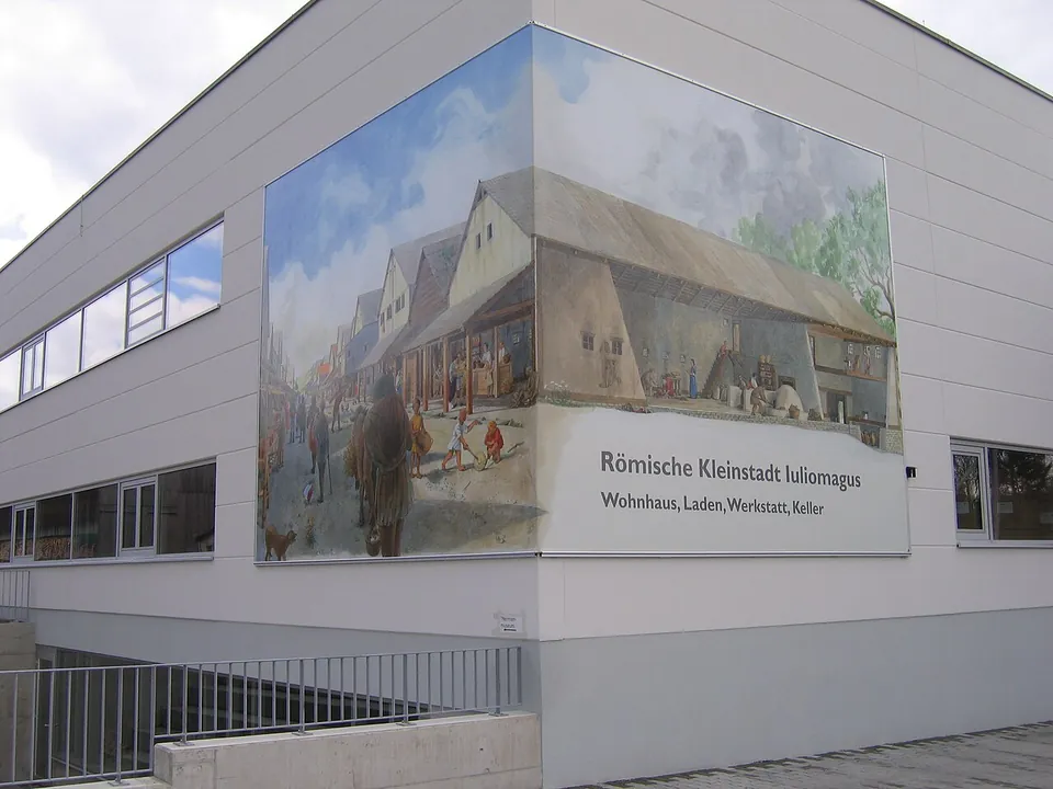 The corner of a modern white commercial building with a large painted panel showing a reconstructed Roman street with timber houses, headed "Römische Kleinstadt Iuliomagus, Wohnhaus, Laden, Werkstatt, Keller".